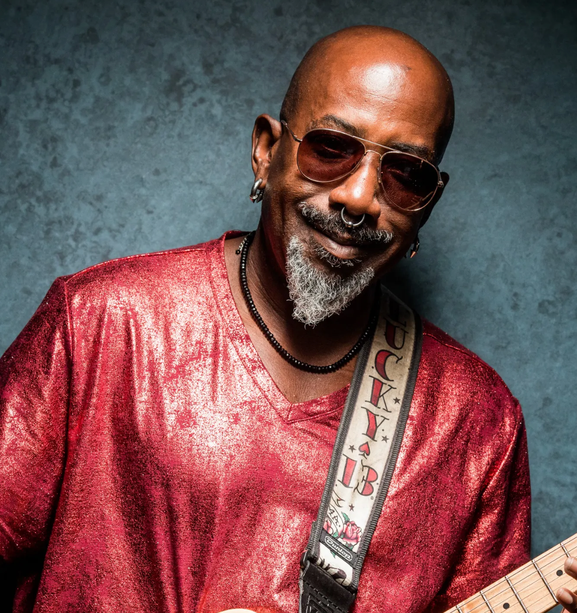 Man wearing sunglasses and a red shirt with a guitar strap over his shoulder 